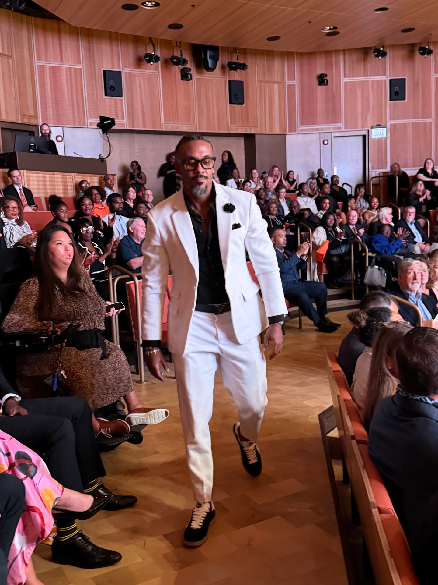 An honoree in a white suit walking down the aisle as the audience applauds