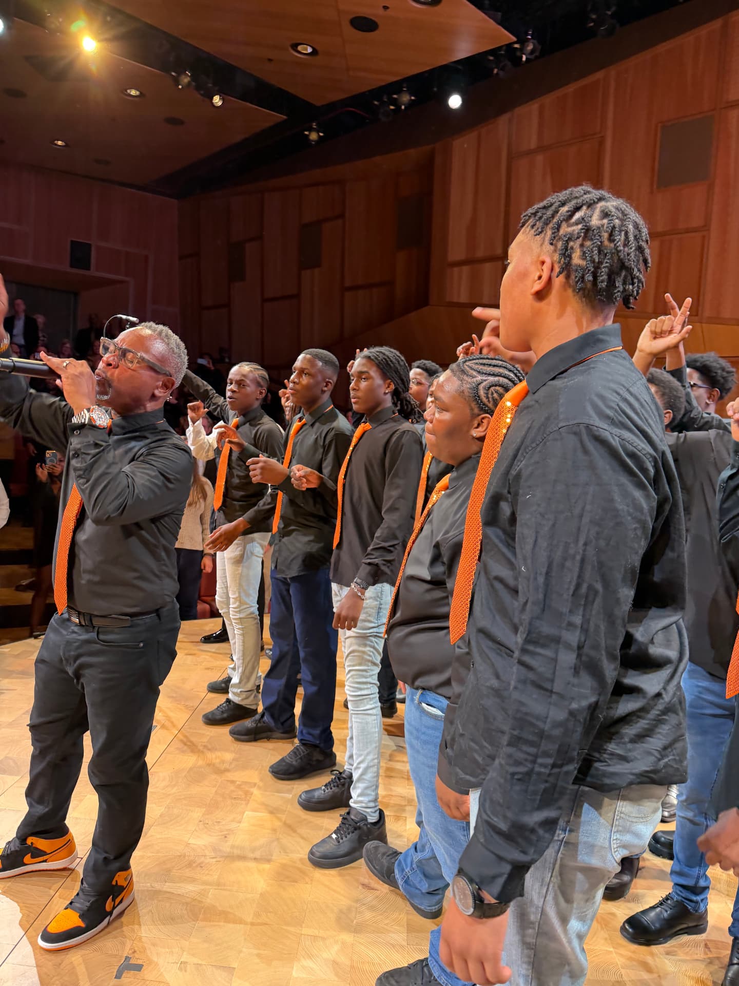 The choir director singing into the microphone beside the choir in orange ties
