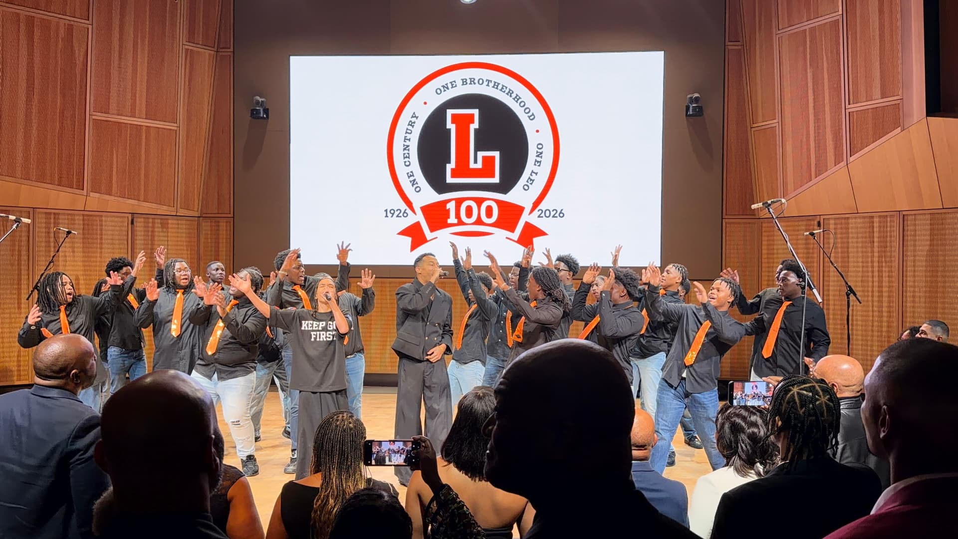The choir raising their hands behind two guest vocalists beneath the Leo 100 crest