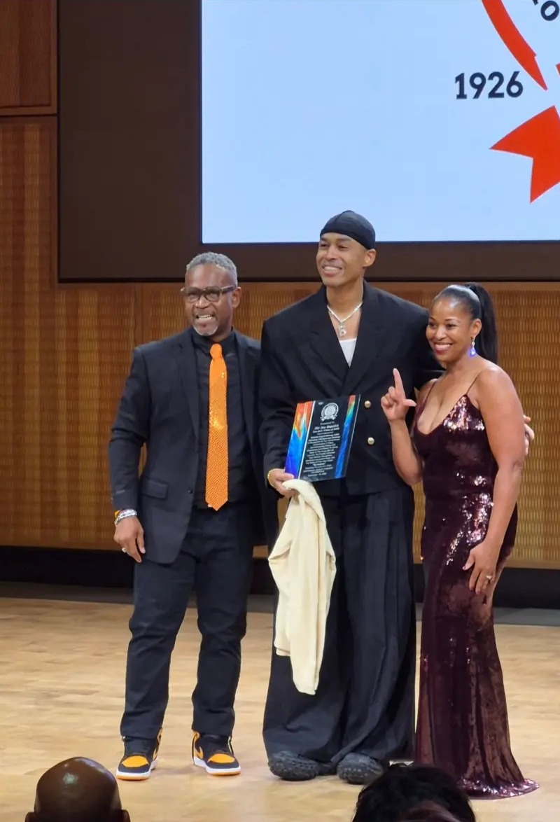 Sir the Baptist holding his award on stage between two presenters
