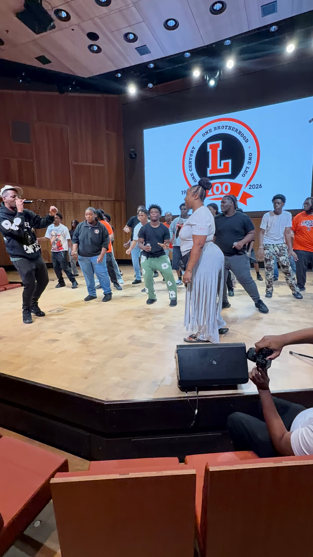 The choir rehearsing on stage in casual clothes beneath the Leo 100 crest