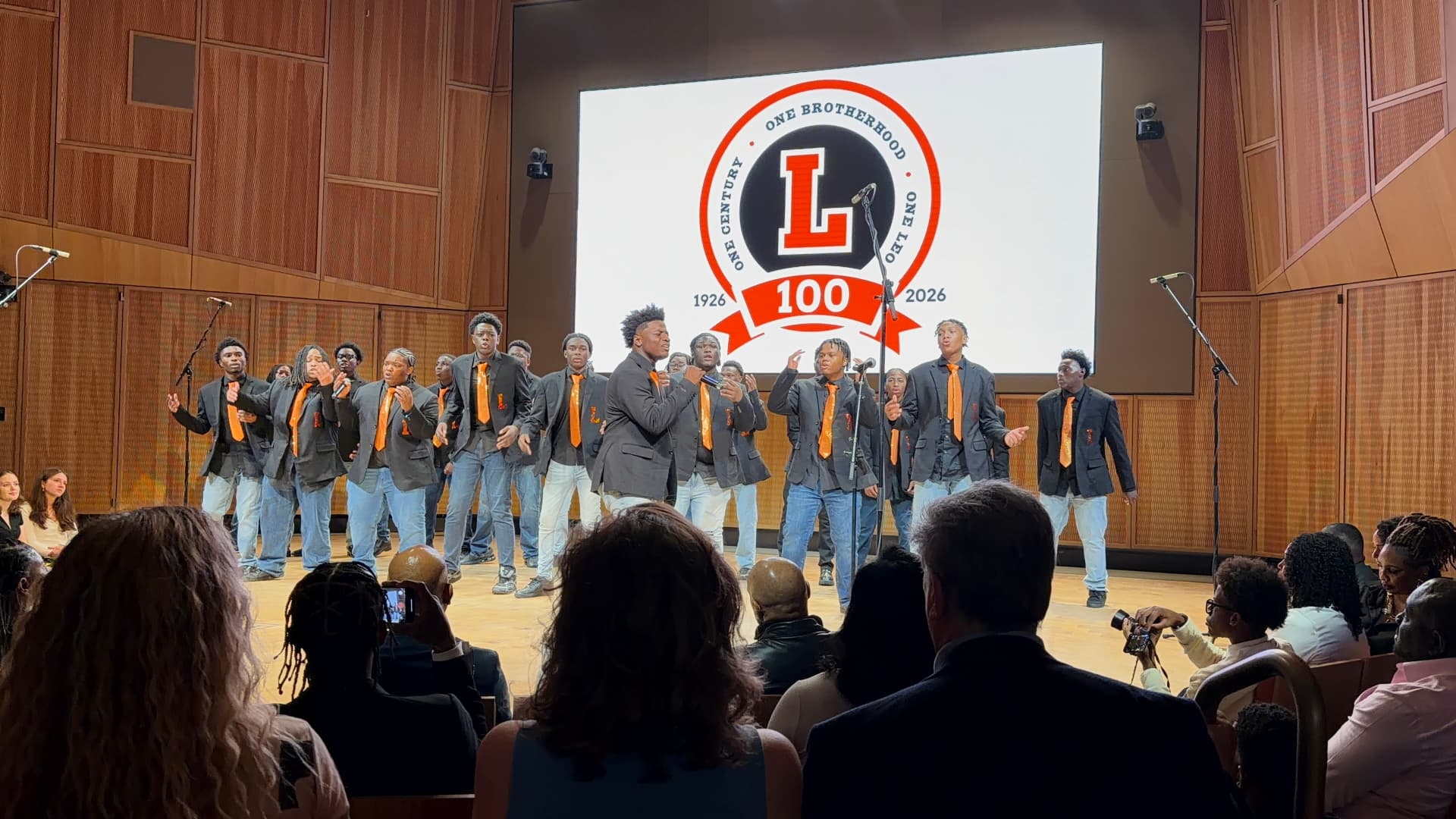 Choir members singing into microphones in front of the Leo 100 crest