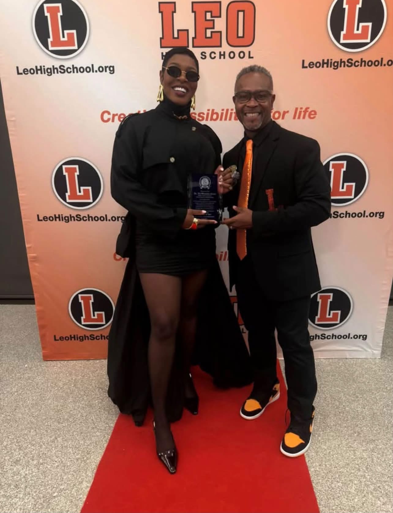 An honoree holding her award with the choir director in front of the Leo backdrop