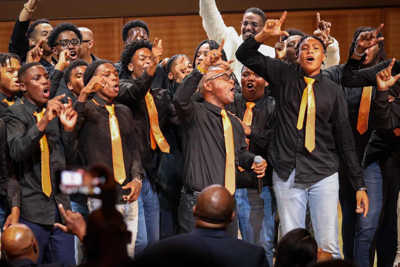 Choir members in gold ties singing with arms raised