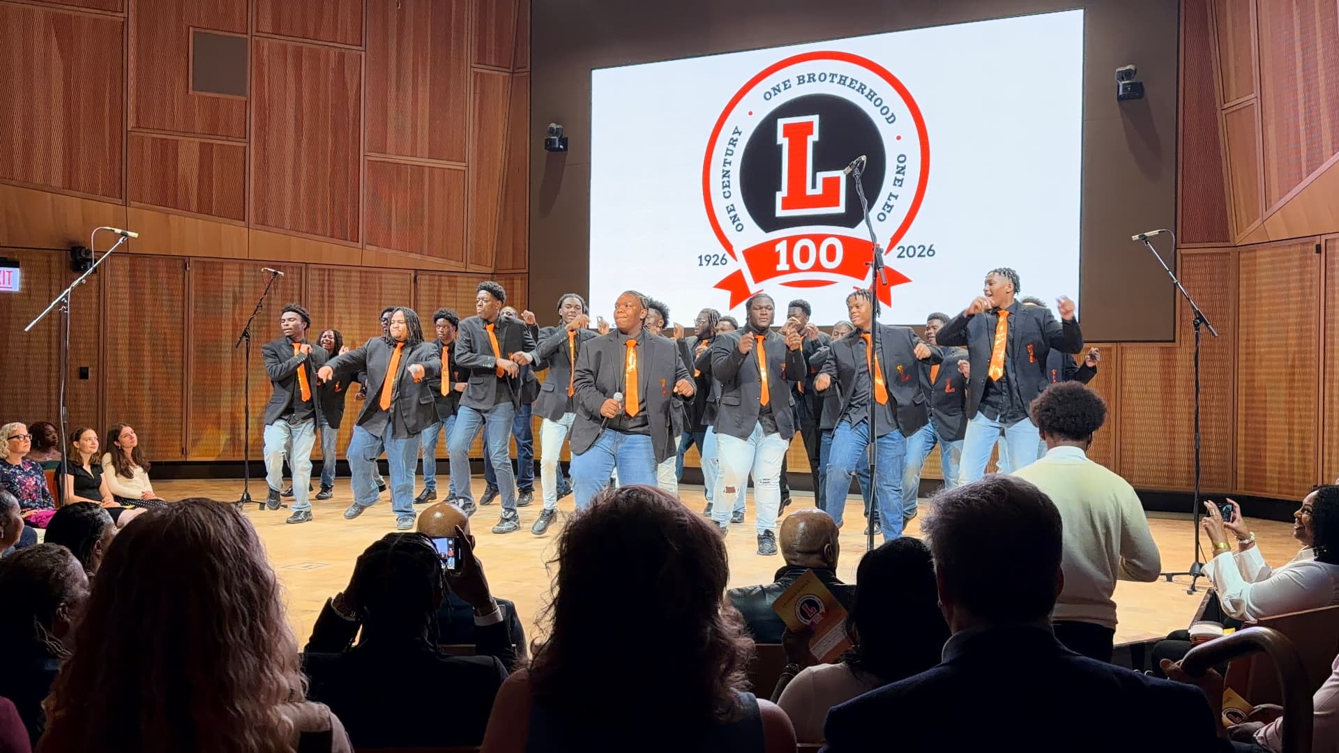 The choir stepping in unison across the stage in blazers and orange ties