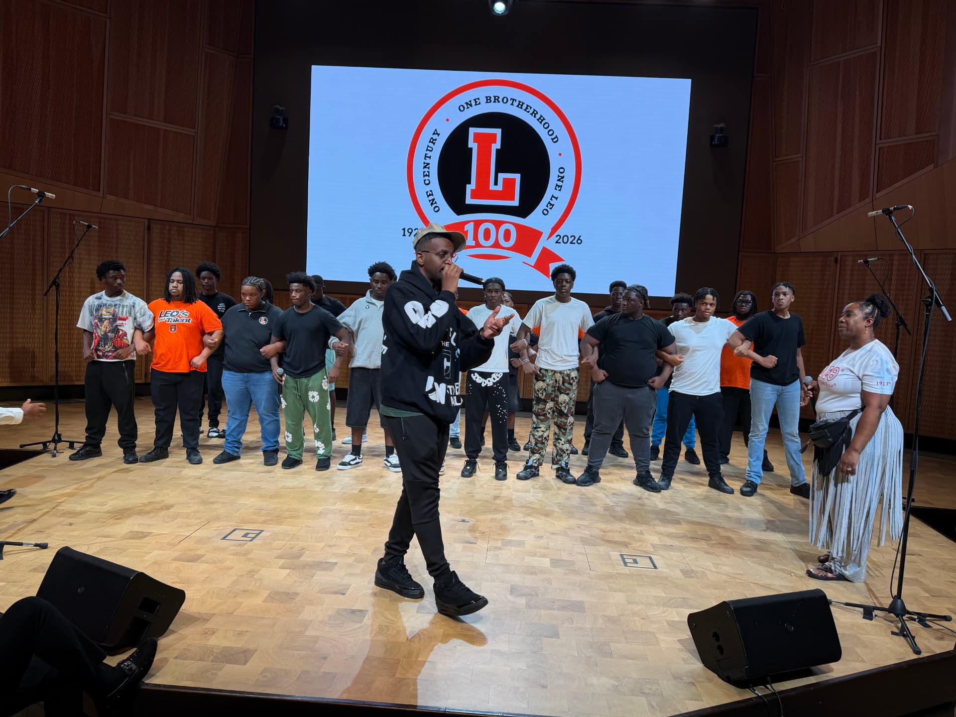 A guest vocalist coaching the choir during rehearsal beneath the Leo 100 crest