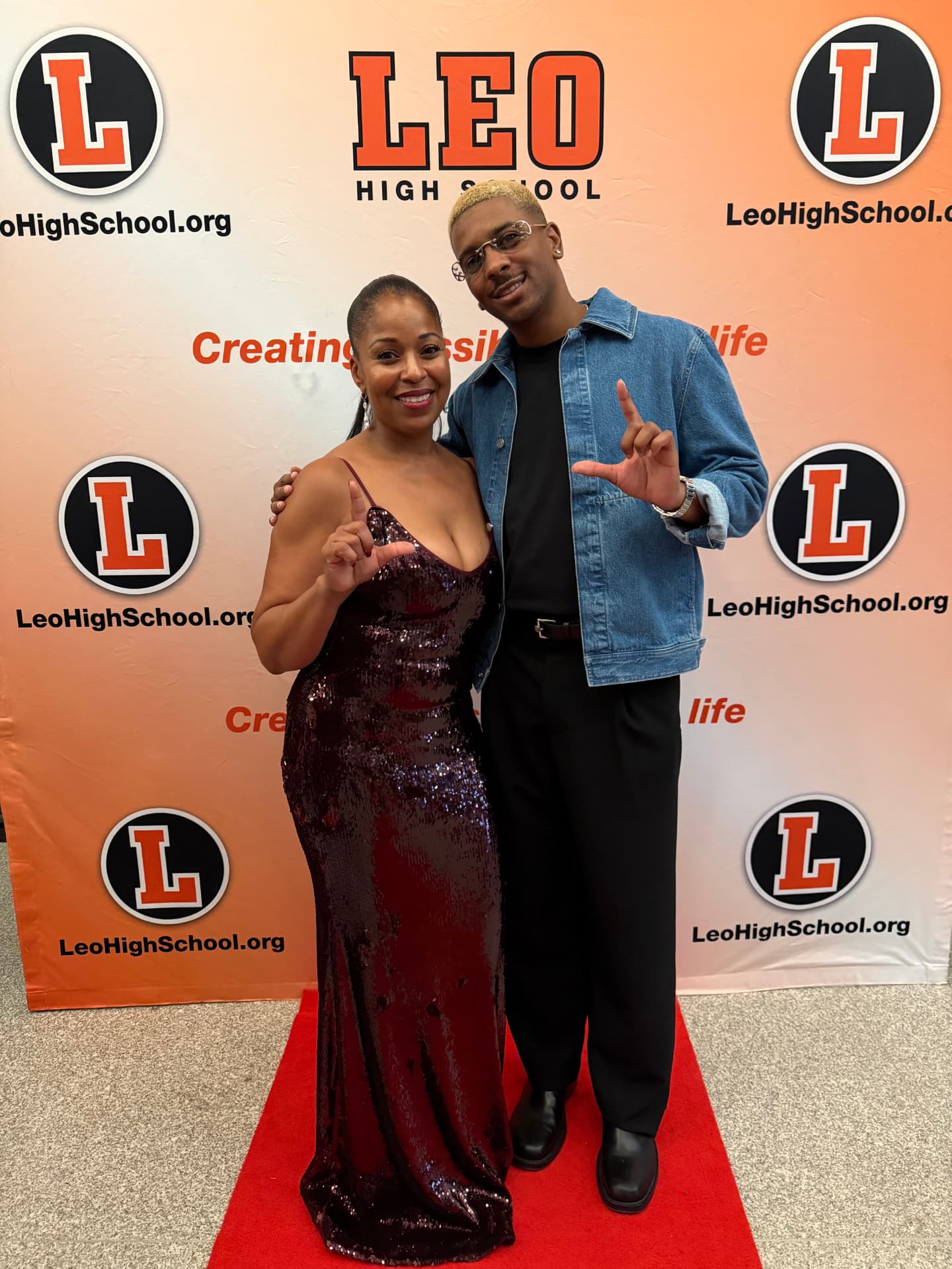 Two guests throwing up the L in front of the Leo High School backdrop