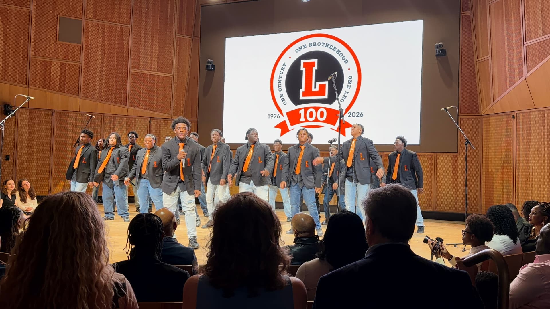 The Leo Choir in orange ties performing in front of the Leo 100 centennial crest