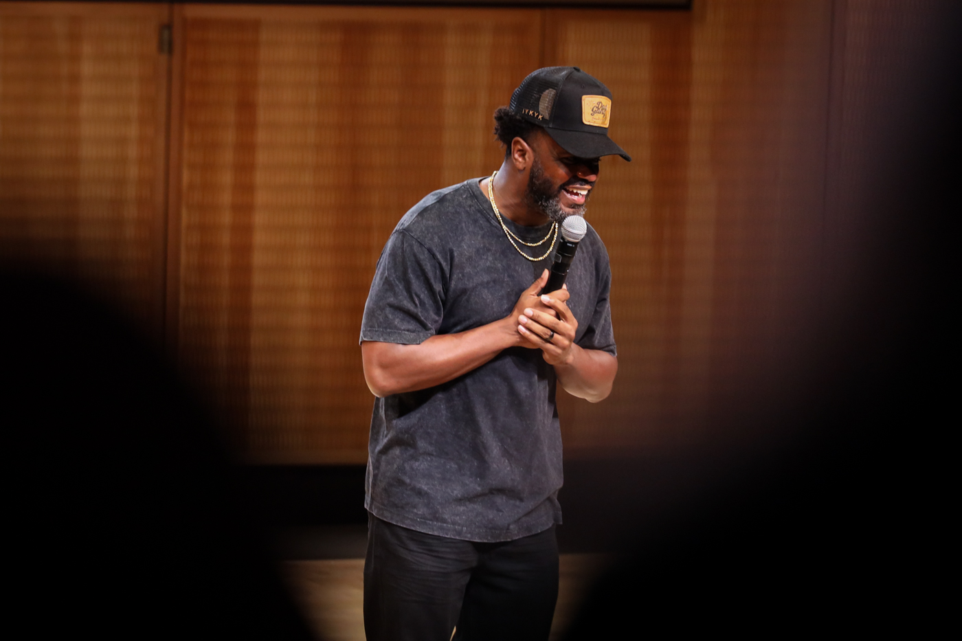 A performer in a black cap smiling as he speaks into the microphone