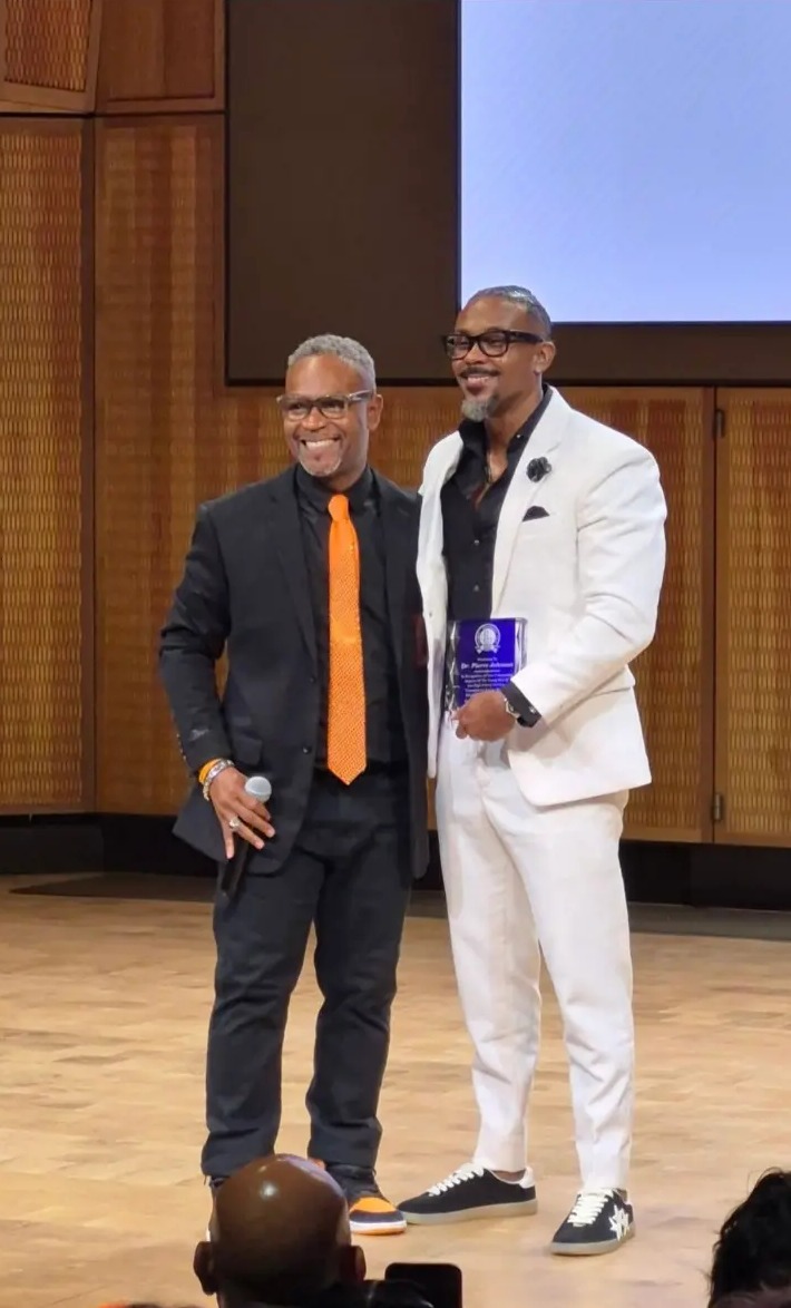 An honoree in a white suit holding his award beside the choir director
