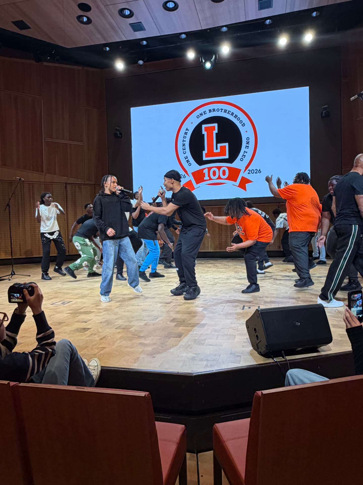 Guest artists with microphones rehearsing with the choir beneath the Leo 100 crest