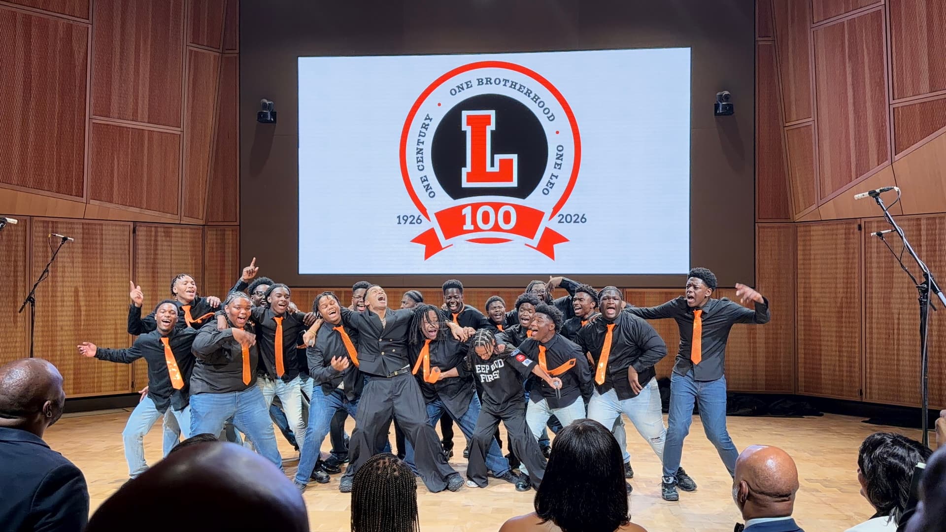 The choir linked arm in arm, laughing and singing with the guest vocalists beneath the Leo 100 crest