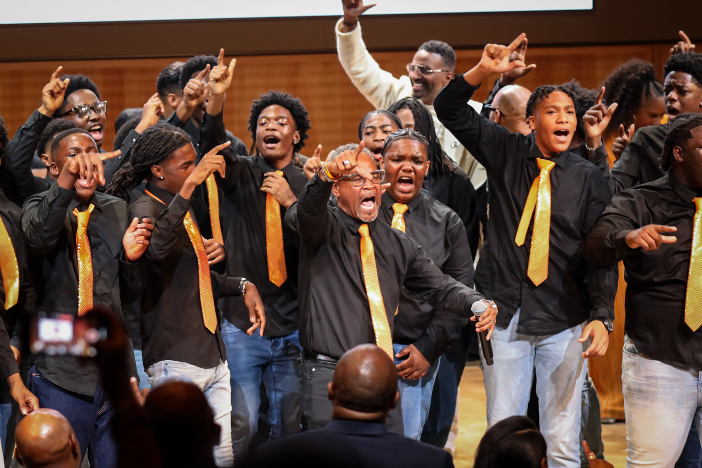 The Leo Choir in gold ties singing with fingers raised in the L sign